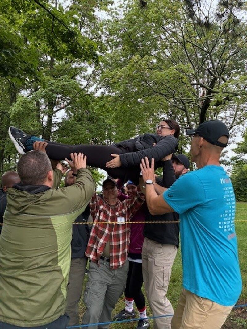 In this image, a group of people are participating in an outdoor team-building activity. One woman is being held up horizontally by several men, who are supporting her with their hands and arms. The participants are dressed casually, and the setting appears to be a park or wooded area with trees in the background. The activity seems to emphasize trust and cooperation among the team members.